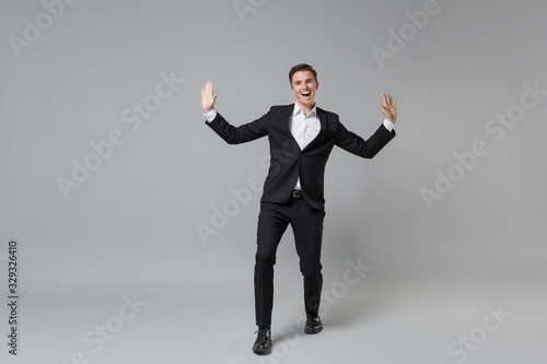 Cheerful laughing young business man in classic black suit shirt posing isolated on grey background studio portrait. Achievement career wealth business concept. Mock up copy space. Spreading hands.