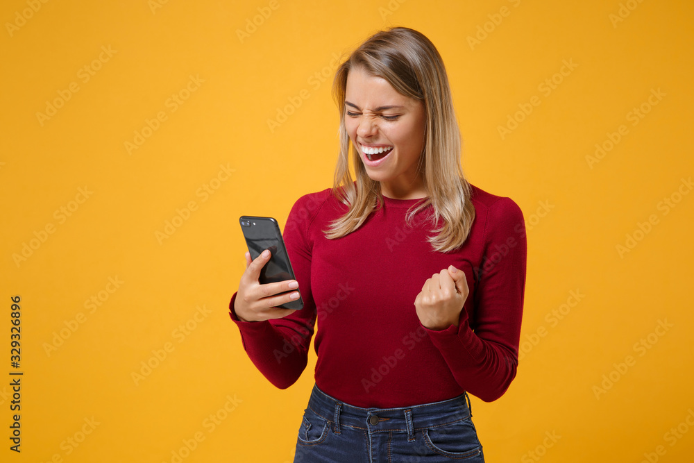 Joyful young blonde woman girl in casual clothes posing isolated on yellow orange background. People lifestyle concept. Mock up copy space. Using mobile phone typing sms message, doing winner gesture. Joyful young blonde woman girl in casual clothes posing isolated on yellow orange background. People lifestyle concept. Mock up copy space. Using mobile phone typing sms message, doing winner gesture.