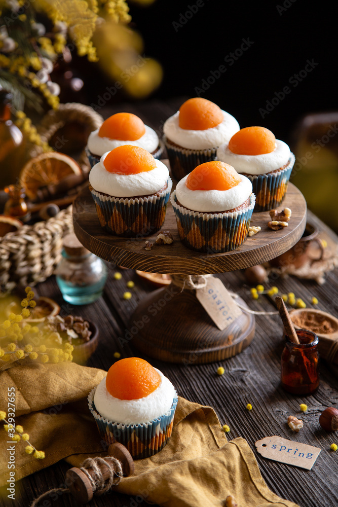 easter carrot cupcakes with white cream and slice of apricot on top (egg style decorated cupcakes) on wooden cake stand on rustic table with willow, oranges, nuts, spices easter carrot cupcakes with white cream and slice of apricot on top (egg style decorated cupcakes) on wooden cake stand on rustic table with willow, oranges, nuts, spices