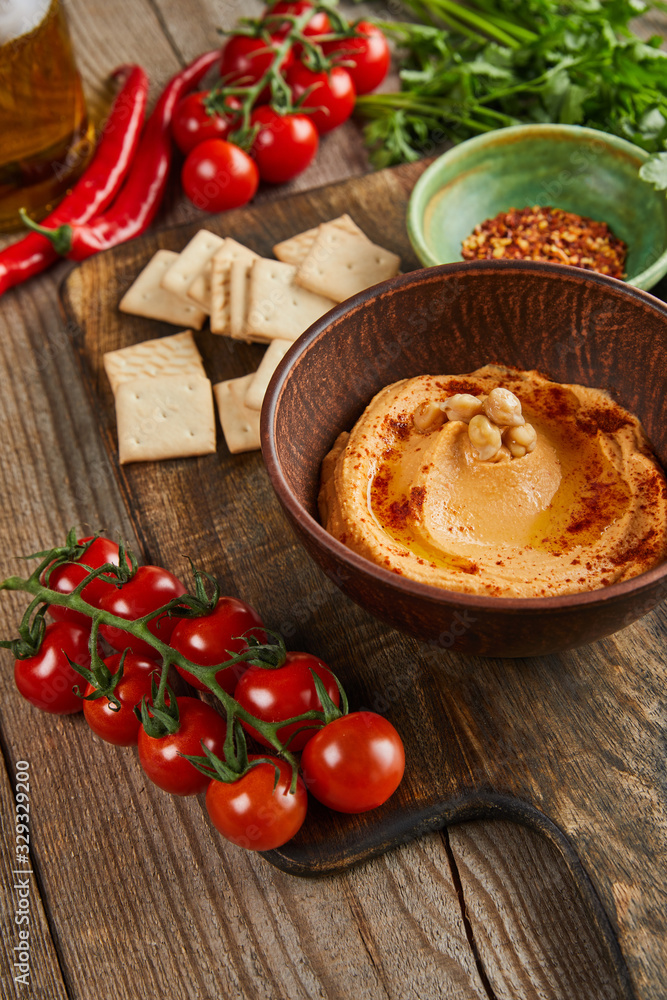Selective focus of bowls with hummus and spices, ripe vegetables and crackers on wooden background Selective focus of bowls with hummus and spices, ripe vegetables and crackers on wooden background
