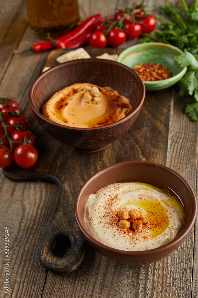 Selective focus of bowls with paprika and hummus, vegetables and parsley on wooden background Selective focus of bowls with paprika and hummus, vegetables and parsley on wooden background