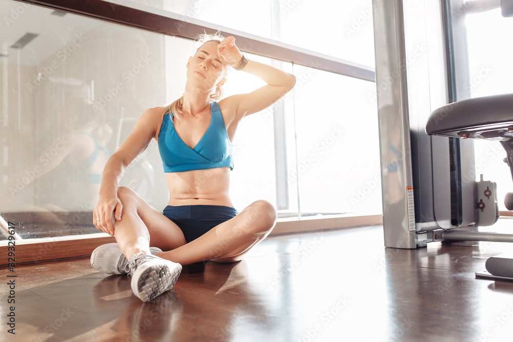 Charming tired purposeful young beautiful caucasian woman in a tracksuit is resting sitting on the floor of the gym after an intensive training. The concept of sports and fitness. Sunset Charming tired purposeful young beautiful caucasian woman in a tracksuit is resting sitting on the floor of the gym after an intensive training. The concept of sports and fitness. Sunset