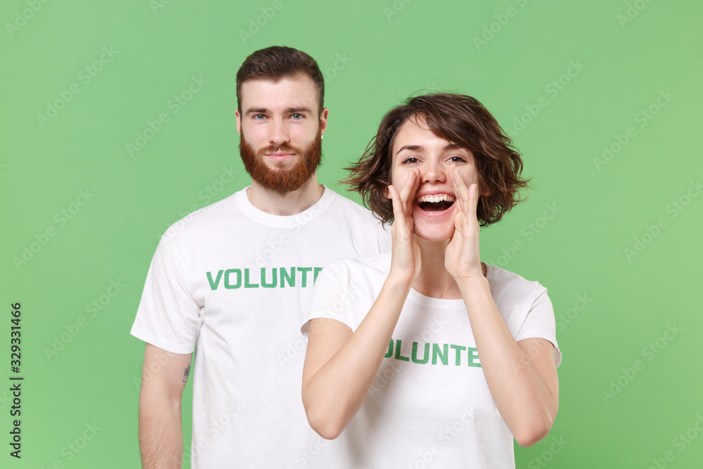 Funny two friends couple in white volunteer t-shirt isolated on pastel green background. Voluntary free work assistance help charity grace teamwork concept. Screaming with hands gesture near mouth. Funny two friends couple in white volunteer t-shirt isolated on pastel green background. Voluntary free work assistance help charity grace teamwork concept. Screaming with hands gesture near mouth.