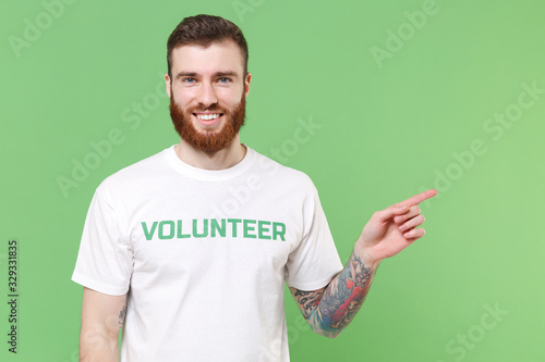 Smiling young bearded man in white volunteer t-shirt isolated on pastel green background studio portrait. Voluntary free work assistance help charity grace teamwork concept. Point index finger aside.