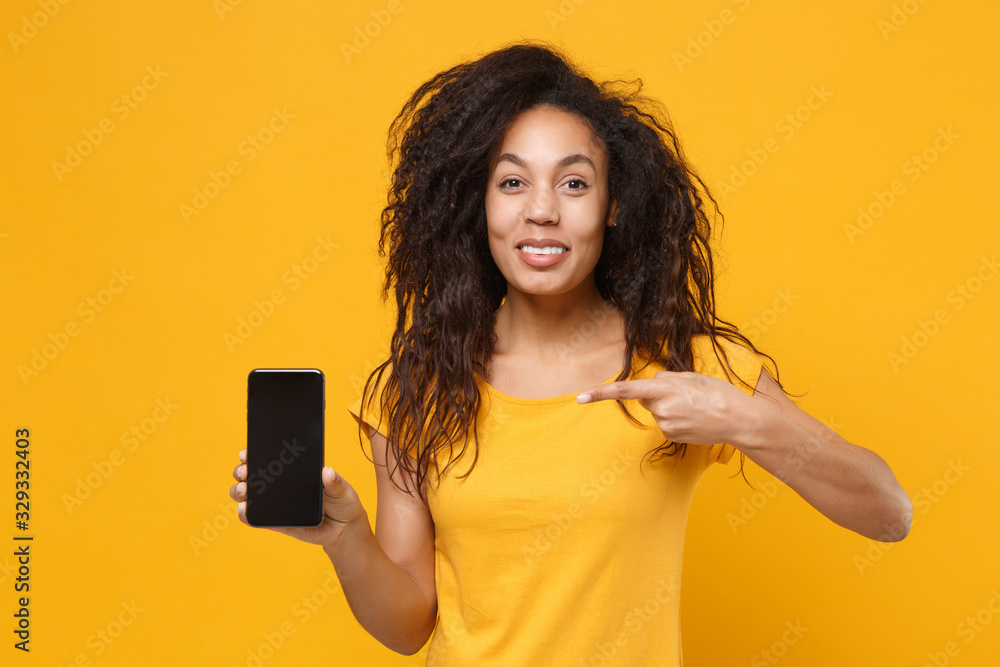 Smiling young african american woman in casual t-shirt isolated on yellow orange background. People lifestyle concept. Mock up copy space. Point index finger on mobile phone with blank empty screen. Smiling young african american woman in casual t-shirt isolated on yellow orange background. People lifestyle concept. Mock up copy space. Point index finger on mobile phone with blank empty screen.