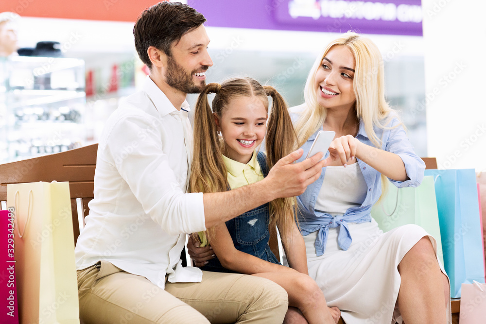 Family Using Smartphone Sitting With Bags On Bench In Mall