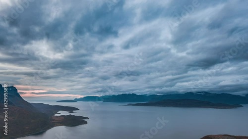 The thick blanket of gray-blue clouds rolling above dark waters of Kvaenangen fjord, Norway. The pale pink light of dawn on the horizon. Dark silhouettes of the mountains framing the picture. photo