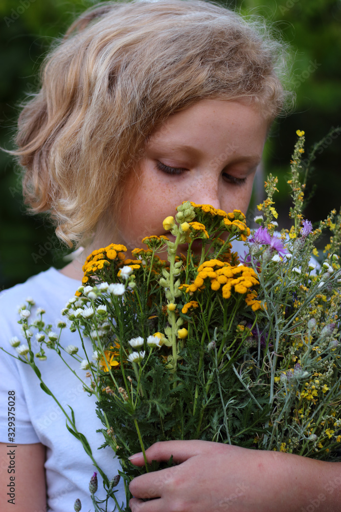 A portrait of the young girl with freckles and blond curly hair. Teen girl holds in her hands a bouquet with wild flowers. Summer. July. Green background. A portrait of the young girl with freckles and blond curly hair. Teen girl holds in her hands a bouquet with wild flowers. Summer. July. Green background.