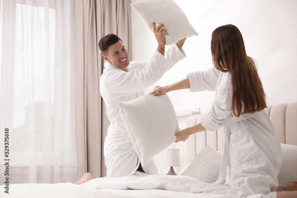 Happy couple in bathrobes having pillow fight on bed at home Happy couple in bathrobes having pillow fight on bed at home