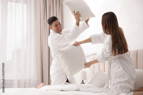 Happy couple in bathrobes having pillow fight on bed at home