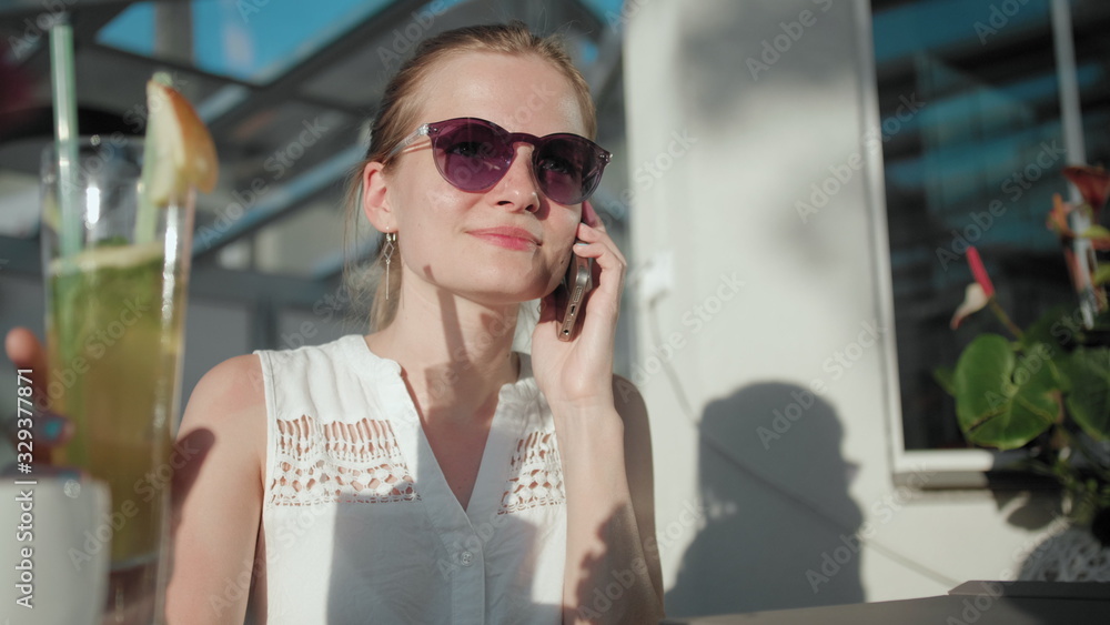Slender Young Adult Woman with Ponytail and Sunglasses is Talking by Phone and Drinking Ice Tea Cocktail with Straw in the Terrace of Cafe or Restaurant in Summer