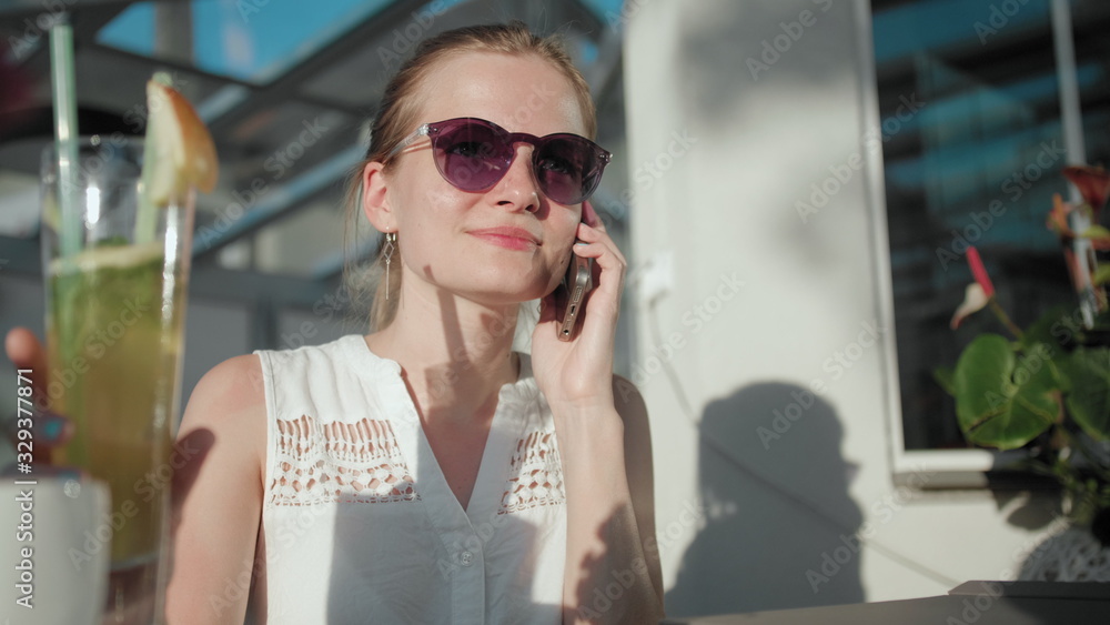 Fototapeta premium Slender Young Adult Woman with Ponytail and Sunglasses is Talking by Phone and Drinking Ice Tea Cocktail with Straw in the Terrace of Cafe or Restaurant in Summer