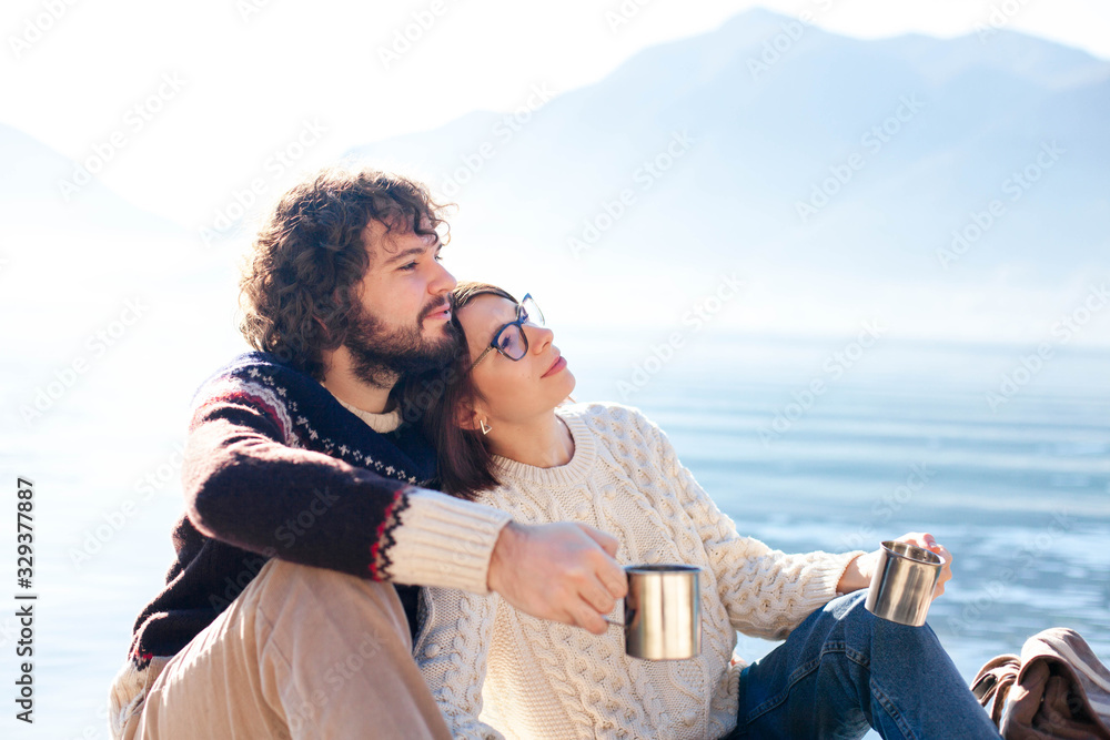 Couple in love drink coffee. Picnic at winter sea beach. Young travelers sitting outdoors. Happy man and woman enjoying nature, relaxation, togetherness and traveling. Romantic lifestyle moment.