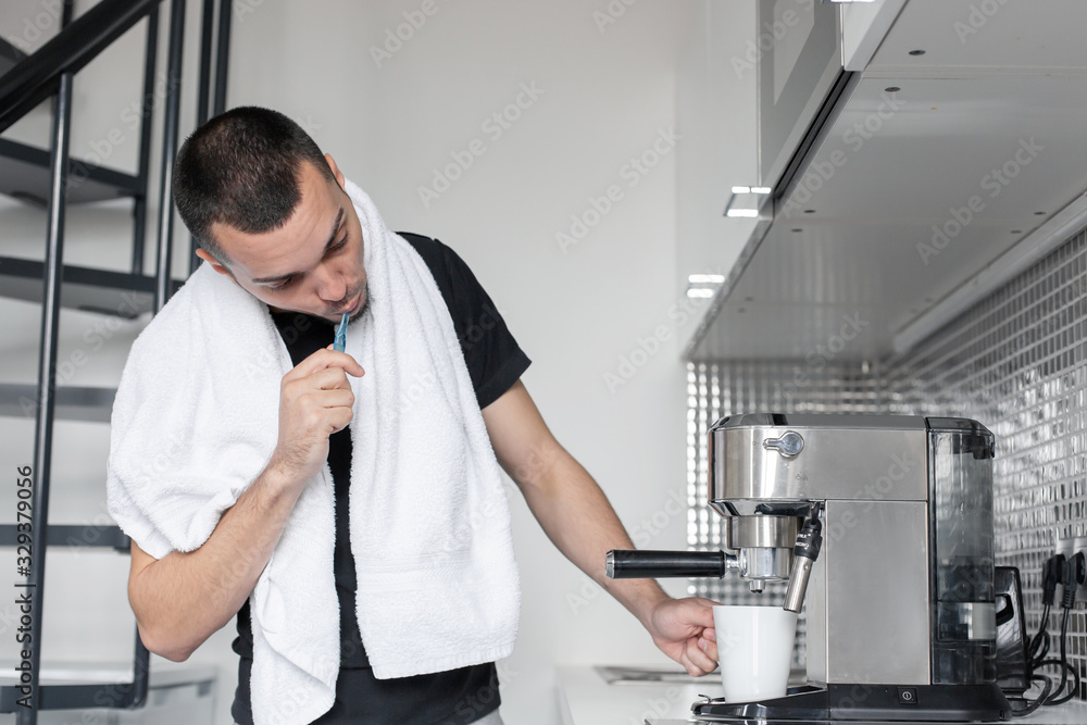 A young guy is going to work in the morning. Brushes teeth near a coffee machine while waiting for a cup of coffee