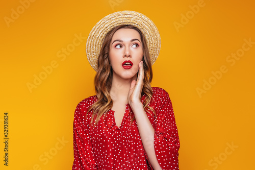 Surprised young girl in a straw hat, in a red dress with a red lipstick looks away at copy space, portrait of a shocked young woman in summer clothes on an orange background, sale concept