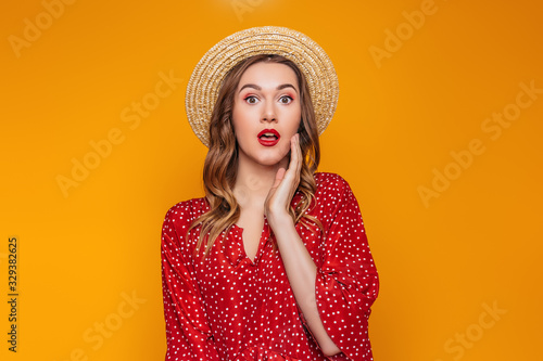 Surprised, shocked young woman in a straw hat, in a red summer dress, red lipstick looks directly at the camera. Portrait of a shocked girl in summer clothes on an orange background, sale concept