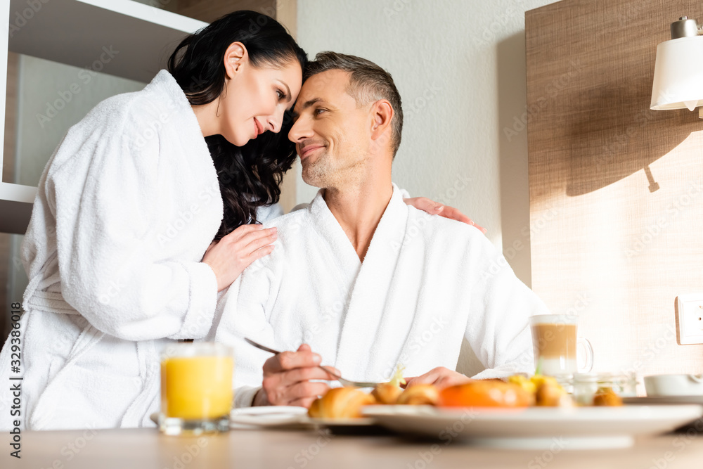 selective focus of smiling girlfriend in bathrobe hugging boyfriend during breakfast