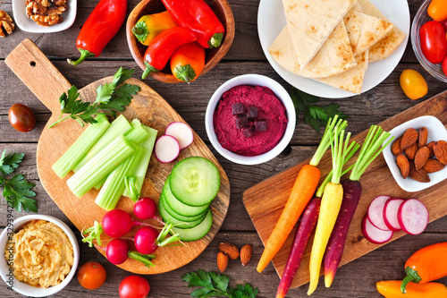 Table scene with a variety of fresh vegetables and hummus dips. Overhead view on a rustic wood background.