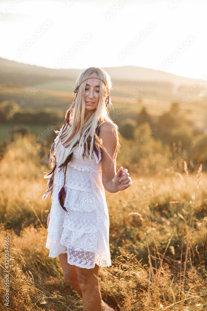 Happy attractive blond boho woman in white dress and feather accessories in hair in summer field outdoors, sunset. Boho fashion, hippie style Happy attractive blond boho woman in white dress and feather accessories in hair in summer field outdoors, sunset. Boho fashion, hippie style