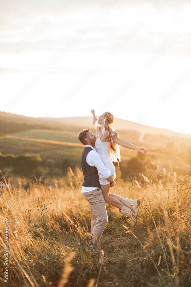 Fototapeta premium Young couple wearing hipster boho indie style clothes, in love walking in countryside, man holds woman on hands, woman is happy and smiling. Sunny sunset field on background, warm summer day