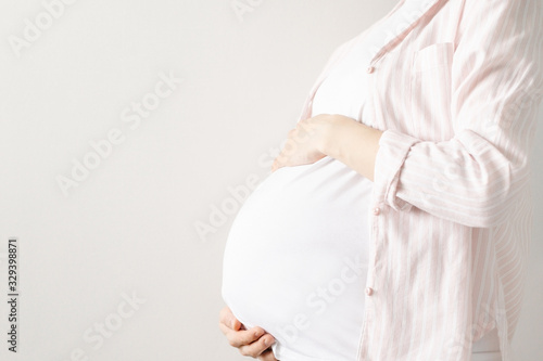 Pregnant woman in a white t-shirt holds her belly with her hands on a light background with copyspace holds her belly with hands. Motherhood and love
