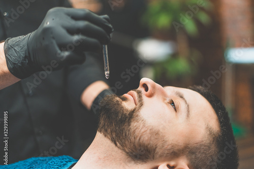 Barbershop. The client in the master’s chair in the barbershop, the barber applies oil and cosmetics to the client’s beard. Male beauty shop. Healthy lifestyle and beauty.