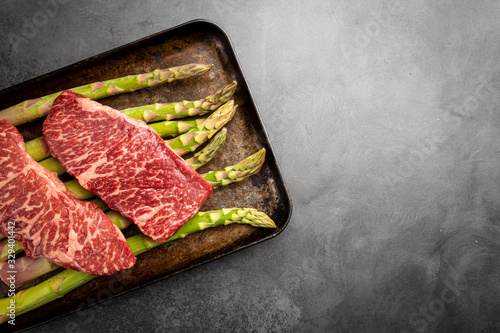 Raw Steak with green asparagus on tray on black background, top view