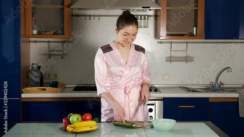 pretty girl in pink dressing-gown makes salad standing at kitchen table and cutting fresh vegetables closeup photo