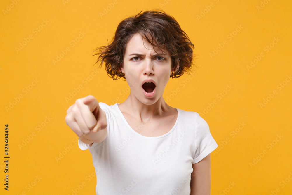 Angry young brunette woman girl in white t-shirt posing isolated on yellow orange background in studio. People lifestyle concept. Mock up copy space. Point index finger on camera swearing screaming. Angry young brunette woman girl in white t-shirt posing isolated on yellow orange background in studio. People lifestyle concept. Mock up copy space. Point index finger on camera swearing screaming.