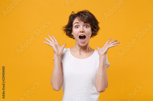 Excited young brunette woman girl in white t-shirt posing isolated on yellow orange background in studio. People emotions lifestyle concept. Mock up copy space. Keeping mouth open, spreading hands.