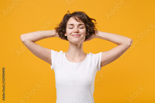 Smiling young brunette woman girl in white t-shirt posing isolated on yellow orange wall background studio portrait. People lifestyle concept. Mock up copy space. Sleeping with hands behind head.
