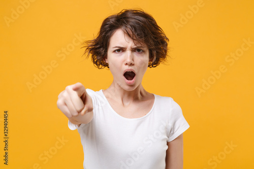 Angry young brunette woman girl in white t-shirt posing isolated on yellow orange background in studio. People lifestyle concept. Mock up copy space. Point index finger on camera swearing screaming.