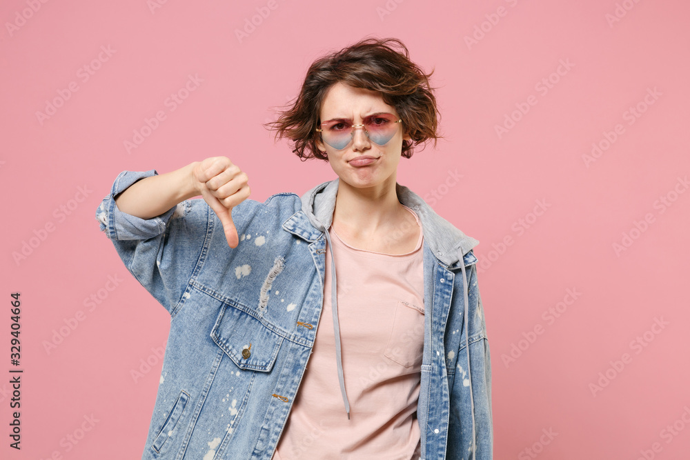 Displeased young brunette woman girl in casual denim jacket eyeglasses posing isolated on pastel pink wall background studio portrait. People lifestyle concept. Mock up copy space. Showing thumb down. Displeased young brunette woman girl in casual denim jacket eyeglasses posing isolated on pastel pink wall background studio portrait. People lifestyle concept. Mock up copy space. Showing thumb down.