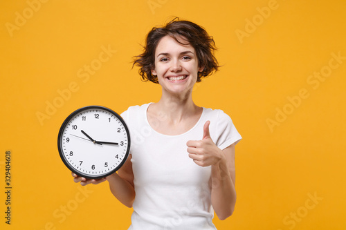 Smiling young brunette woman girl in white t-shirt posing isolated on yellow orange background studio portrait. People emotions lifestyle concept. Mock up copy space. Hold clock, showing thumb up.
