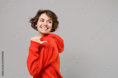 Side view of smiling young brunette woman girl in casual red hoodie posing isolated on grey background studio portrait. People emotions lifestyle concept. Mock up copy space. Pointing thumb aside.