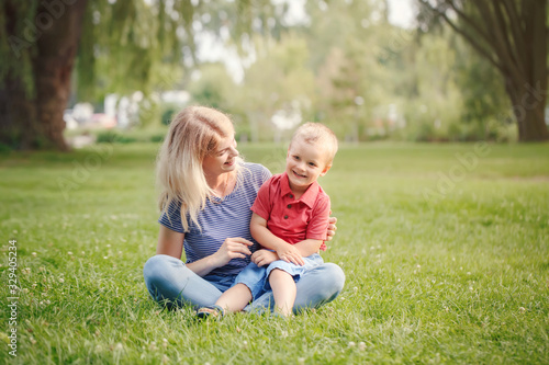 Young smiling Caucasian mother and laughing boy toddler son sitting on grass in park. Family mom and child hugging having fun outdoor on a summer day. Happy authentic family childhood lifestyle.