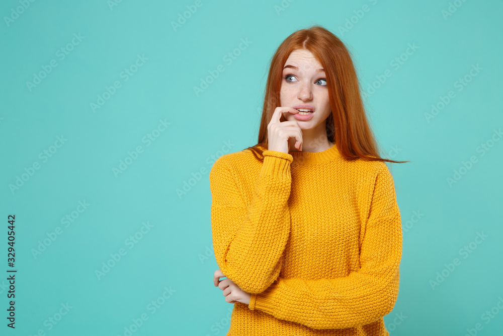 Perplexed pensive young redhead woman girl in yellow sweater posing isolated on blue turquoise background studio portrait. People lifestyle concept. Mock up copy space. Gnawing nails, looking aside. Perplexed pensive young redhead woman girl in yellow sweater posing isolated on blue turquoise background studio portrait. People lifestyle concept. Mock up copy space. Gnawing nails, looking aside.