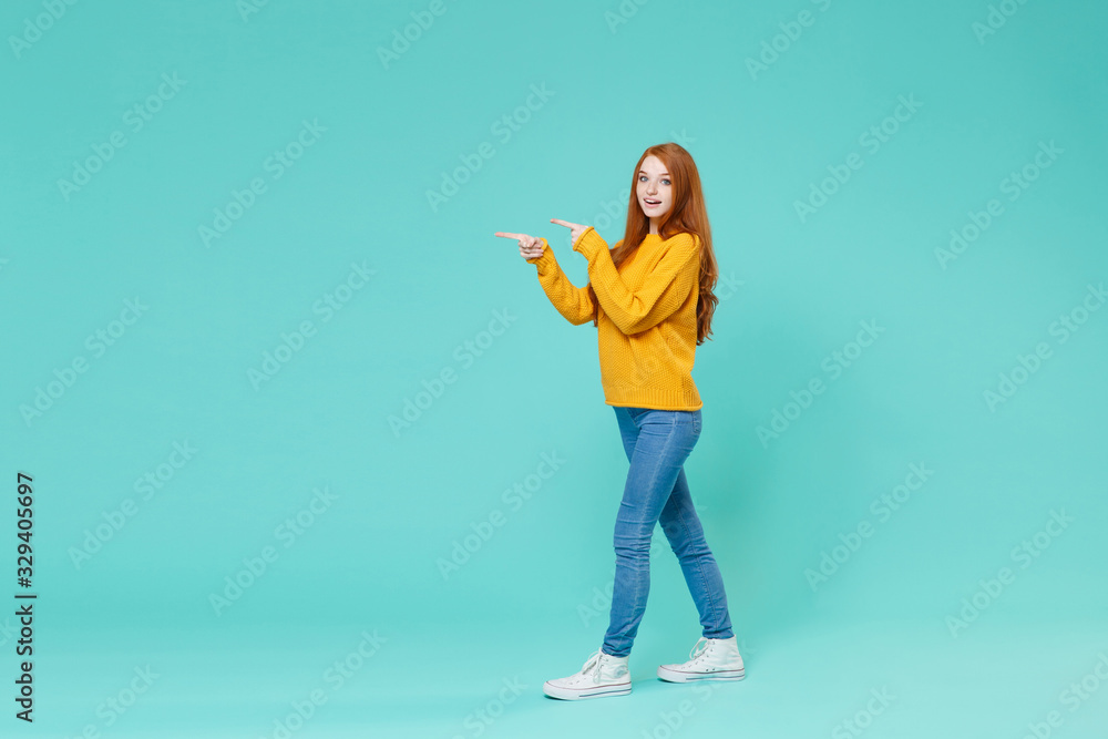 Pretty young redhead woman girl in yellow sweater posing isolated on blue turquoise background studio portrait. People emotions lifestyle concept. Mock up copy space. Pointing index fingers aside. Pretty young redhead woman girl in yellow sweater posing isolated on blue turquoise background studio portrait. People emotions lifestyle concept. Mock up copy space. Pointing index fingers aside.