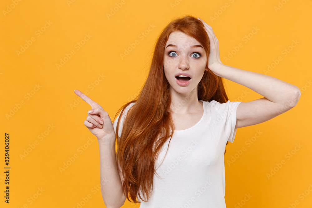 Amazed young redhead woman girl in white blank empty t-shirt posing isolated on yellow background in studio. People lifestyle concept. Mock up copy space. Point index finger aside up put hand on head. Amazed young redhead woman girl in white blank empty t-shirt posing isolated on yellow background in studio. People lifestyle concept. Mock up copy space. Point index finger aside up put hand on head.