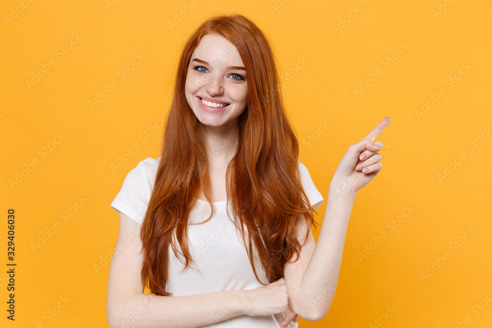 Smiling young redhead woman girl in white blank empty t-shirt posing isolated on yellow wall background studio portrait. People lifestyle concept. Mock up copy space. Pointing index finger aside up. Smiling young redhead woman girl in white blank empty t-shirt posing isolated on yellow wall background studio portrait. People lifestyle concept. Mock up copy space. Pointing index finger aside up.