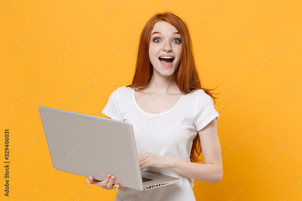 Surprised young redhead woman girl in white blank empty t-shirt posing isolated on yellow wall background studio portrait. People lifestyle concept. Mock up copy space. Working on laptop pc computer. Surprised young redhead woman girl in white blank empty t-shirt posing isolated on yellow wall background studio portrait. People lifestyle concept. Mock up copy space. Working on laptop pc computer.