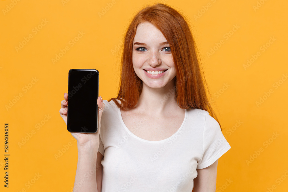 Smiling young redhead woman girl in white t-shirt posing isolated on yellow wall background studio portrait. People lifestyle concept. Mock up copy space. Hold mobile phone with blank empty screen. Smiling young redhead woman girl in white t-shirt posing isolated on yellow wall background studio portrait. People lifestyle concept. Mock up copy space. Hold mobile phone with blank empty screen.