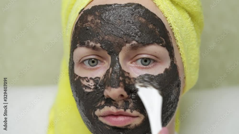 A girl with a towel on her head puts purifying black mask on her face by a cosmetic spatula. Charcoal face mask or black clay mud. Natural cosmetic concept. Skin care, acne treatment, cleansing skin. A girl with a towel on her head puts purifying black mask on her face by a cosmetic spatula. Charcoal face mask or black clay mud. Natural cosmetic concept. Skin care, acne treatment, cleansing skin.