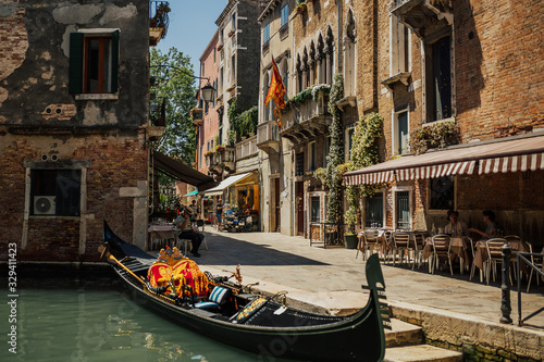 Obraz na płótnie Charming landscape of typical street in Venice. Open-air cafe near canal with emerald water. Gondola parked near cafe. Sunny day. w salonie