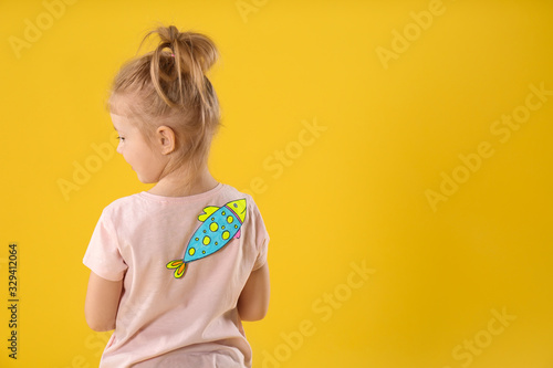 Little girl with paper fish on back against yellow background, space for text. April fool's day