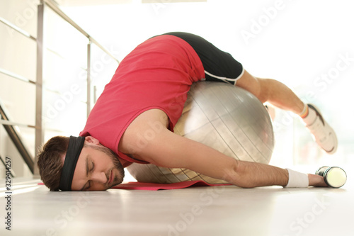 Lazy young man with exercise ball on yoga mat indoors