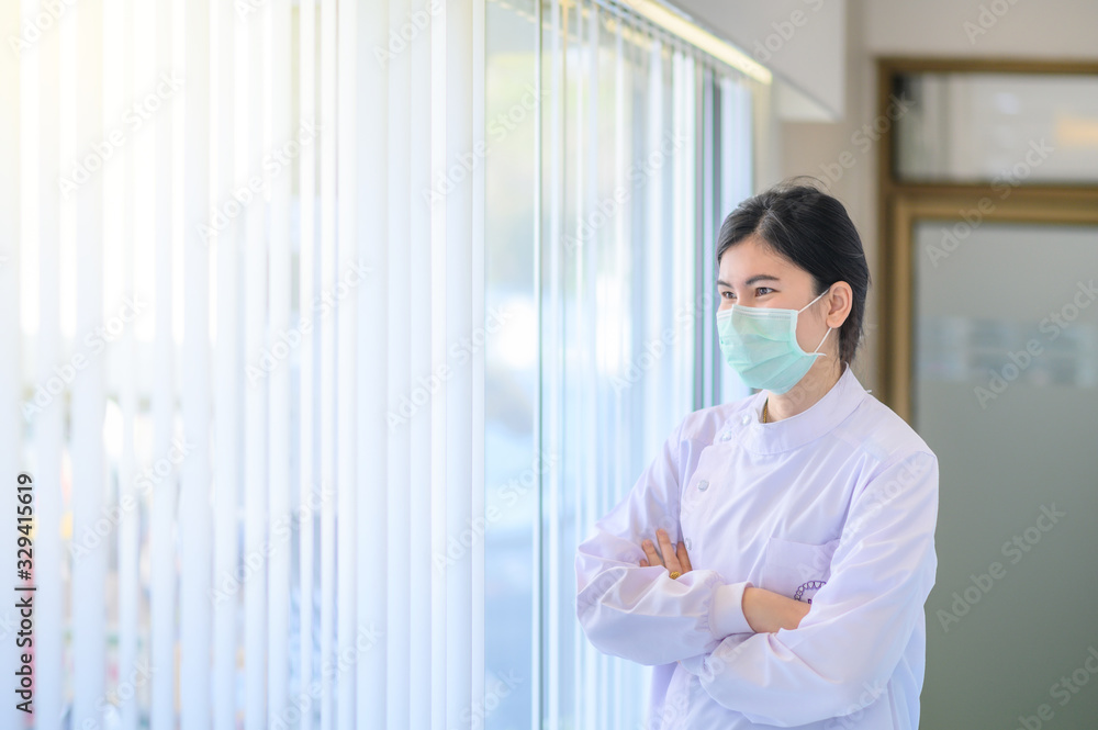 Female dentist wearing surgical mask crossed her arms, standing near the windows and looking out Female dentist wearing surgical mask crossed her arms, standing near the windows and looking out