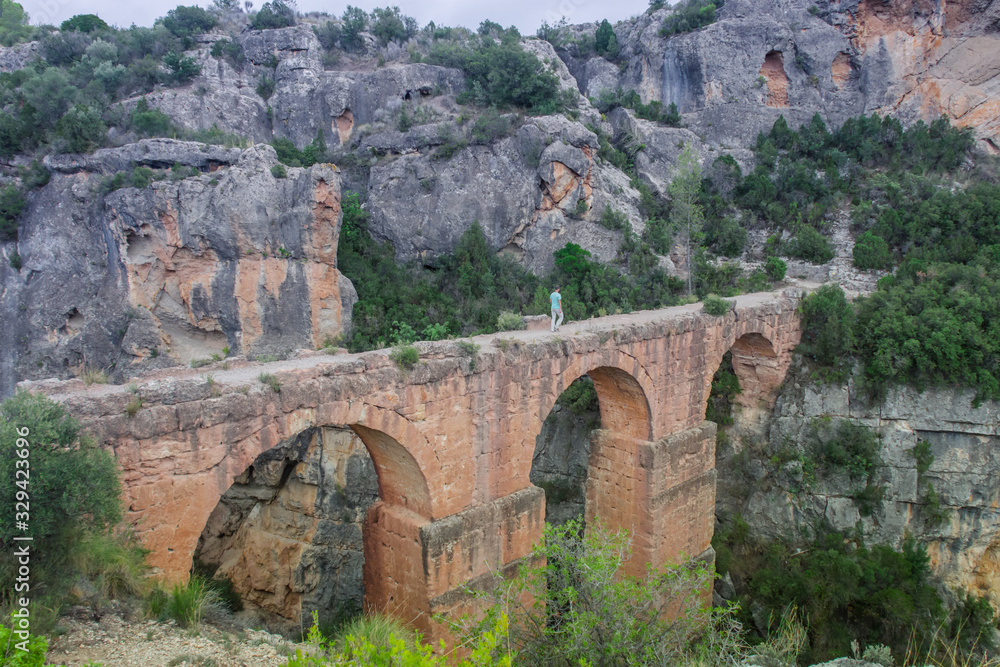 Ancient roman aqueduct with a man walking on it surrounded by rocky mountains in Chelva, valencian community, Spain