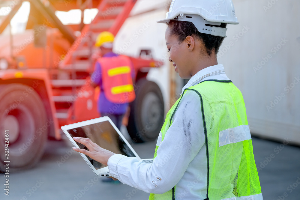 African technician worker woman touch and look to laptop while her co-worker work with construction vehicle.
