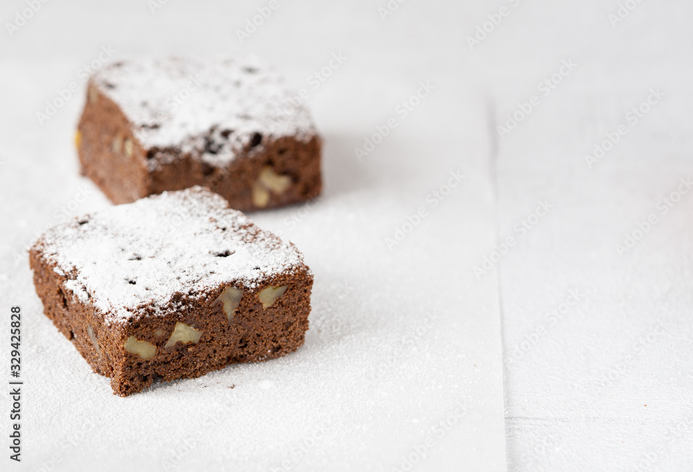 Two homemade chocolate brownie with nuts with icing sugar on white layer of paper. Close up chocolate cake. Copy space. Grey background. Concept: pastry, American traditional dessert, bakery. Two homemade chocolate brownie with nuts with icing sugar on white layer of paper. Close up chocolate cake. Copy space. Grey background. Concept: pastry, American traditional dessert, bakery.